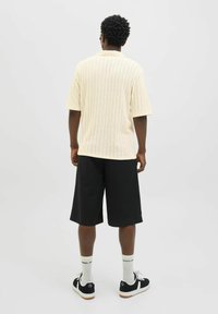 Maglione corto a maniche corte di un beige chiaro con una texture a coste, abbinato a pantaloni corti neri larghi. Le scarpe sono sneakers nere con dettagli bianchi.