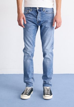 Personne portant un jean bleu clair et des baskets basses noires, debout sur un sol bleu devant un fond blanc.