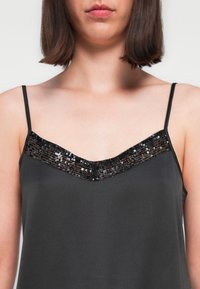 Camisole noire avec un décolleté en V orné d'une bordure en sequins noirs. Fines bretelles ajustables, tissu lisse avec une texture douce.