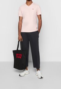 Camiseta de cuello en V rosa claro, bolsa de mano negra con el logo rojo de Levi's, pantalones azul marino y zapatillas blancas. La camiseta presenta un pequeño logo en el pecho.