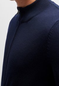 Tummansininen vetoketjullinen neulepaita korkealla kauluksella, valmistettu pehmeästä kankaasta. Siinä on hieno neulos, sileä pinta ja minimalistinen muotoilu.