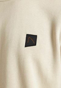 Tissu couleur crème avec un patch noir texturé orné d'un motif de lignes oranges, cousu sur un vêtement. Texture globale lisse.