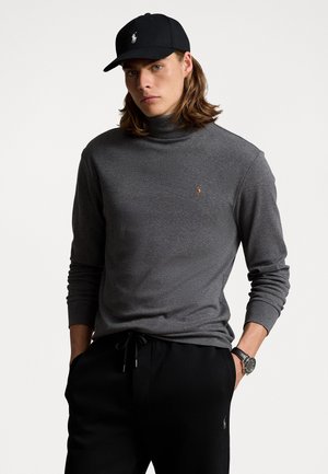 Donkergrijze coltrui van zacht materiaal, met een klein embleem, gecombineerd met zwarte joggingbroeken en een zwarte pet.