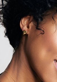 Złota kolczyk w kształcie serca noszona na płatku ucha; gładka, refleksyjna powierzchnia z wypolerowanym wykończeniem, doskonale komponująca się z karnacją.