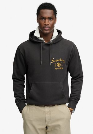 Giovane uomo che indossa una felpa nera Superdry con cappuccio sopra una camicia bianca con colletto, pantaloni beige e un orologio da polso, in piedi con le mani in tasca.