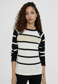 Randig långärmad topp i svart och vitt med gulddetaljer; har en rund halsringning och dekorativa knappar på axeln.