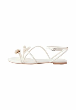 White flat sandal with thin straps, ankle buckle, and small floral decorations near the toes on a white background.