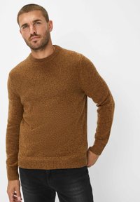 Bruine gebreide trui met een gestructureerd patroon, ronde halslijn en geribbelde manchetten. In combinatie met donkere broek, wat een casual uitstraling geeft.