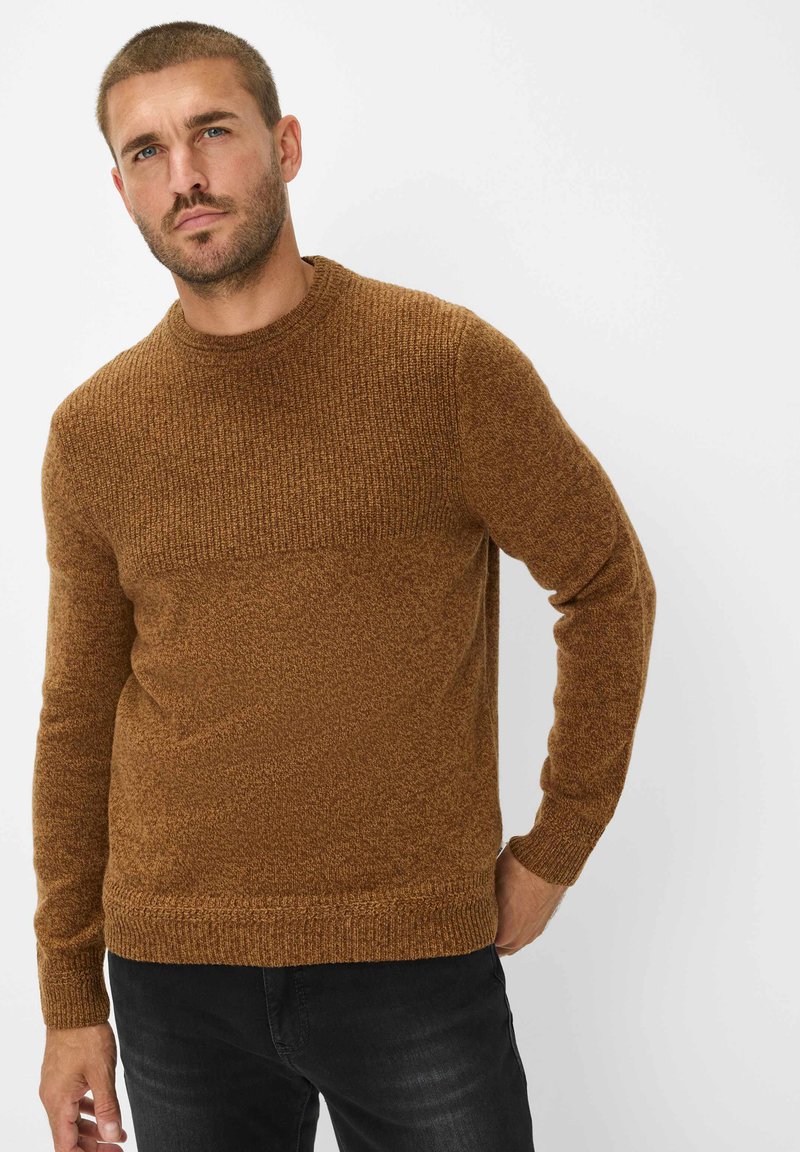 Bruine gebreide trui met een gestructureerd patroon, ronde halslijn en geribbelde manchetten. In combinatie met donkere broek, wat een casual uitstraling geeft.