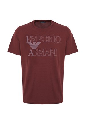 Jeune homme ajustant la manche d'un t-shirt bordeaux Emporio Armani, portant un short blanc et des baskets blanches, montrant des tatouages de serpent et de fleurs sur le bras.