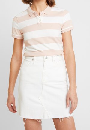 Kobieta w beżowo-białej koszulce polo w paski, włożonej w białą dżinsową spódnicę z przednim rozcięciem i postrzępionym dołem.