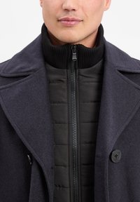 Mann in einem dunklen Wollmantel über einer schwarzen gesteppte Jacke mit einem gerippten hohen Kragen und einem Reißverschluss an der Vorderseite, sichtbar vom Hals bis zur Brust.