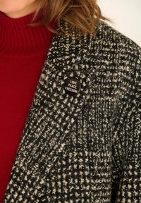 Manteau à carreaux noir et blanc en tissu texturé, avec un col roulé rouge en dessous. Une broche ronde noire affiche le texte "AMOUR. PAIX. LIBERTÉ."