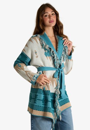 Donna che indossa un cardigan lavorato a maglia con fantasia nei toni turchese, beige e crema con dettagli a frange e cintura annodata, posando con la mano vicino al colletto.