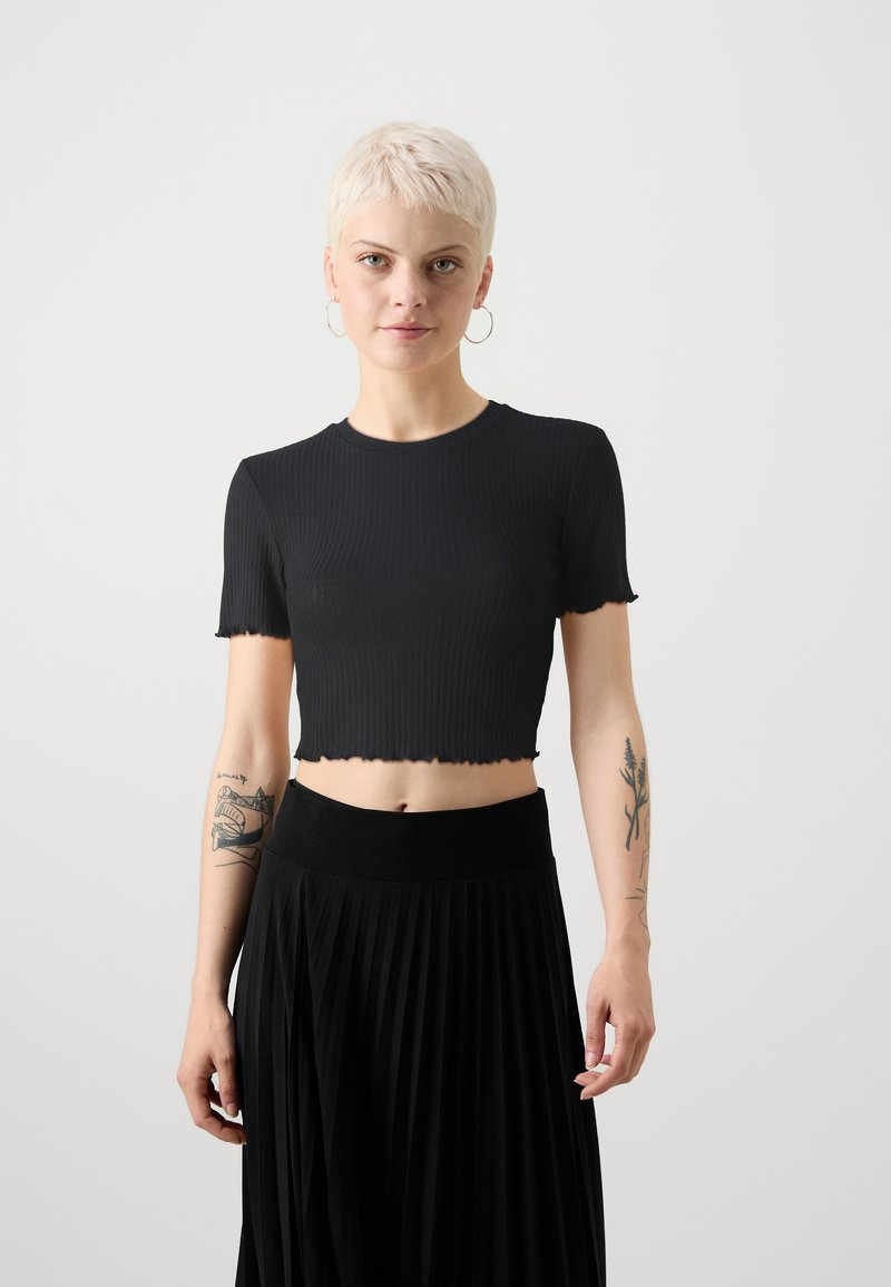 Černý žebrovaný crop top s krátkými rukávy, který má vlnitý lem. Nosen s černou plisovanou sukní s vysokým pasem. Na pažích je viditelný tón pleti.