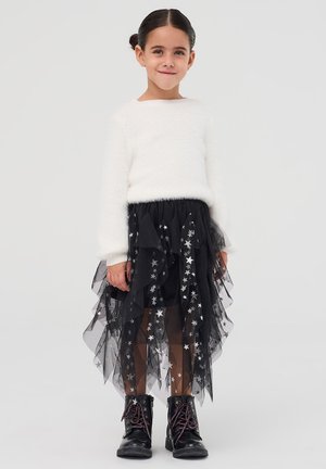 Giovane ragazza con capelli scuri raccolti in un bun, che indossa un maglione bianco peloso, una gonna nera di tulle con motivo a stelle e stivali neri con lacci, in piedi e sorridente.