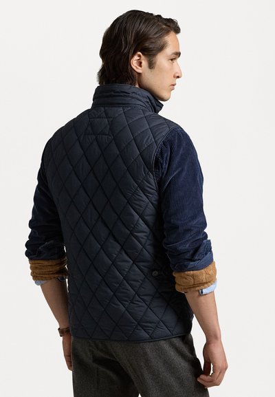 Marineblå quilted vest med høj krave, der har diamantsting og er lagt over en kordfløjlsskjorte med opkørte ærmer.