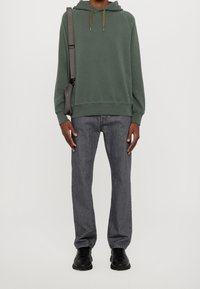 Felpa verde con cappuccio e stringhe grigie, abbinata a jeans grigi e sneaker nere. Il modello porta una borsa a spalla grigia.