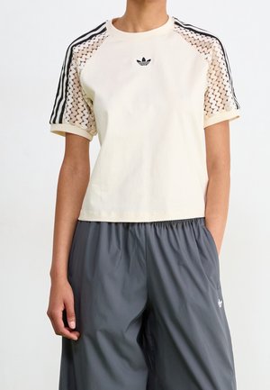 Personne portant un t-shirt crème à manches courtes avec épaules en dentelle et rayures noires, associé à un pantalon gris ample avec ceinture élastique.
