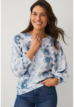 Smilende kvinde med langt brunt hår iført sølvfarvede øreringe, en blå og hvid blomstret sweater og blå jeans, stående mod en lys baggrund.