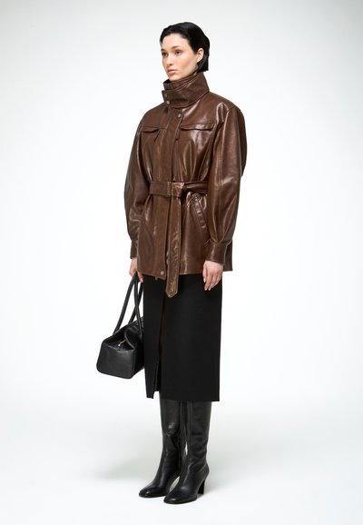 Femme portant une veste en cuir marron ceinturée, une jupe midi noire, des bottes noires montantes jusqu'aux genoux, et tenant un sac à main noir devant un fond uni.