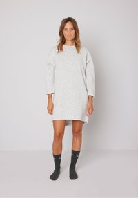 Šviesiai pilka, per didelė megztinio suknelė su ilgomis rankovėmis, šoninėmis skaidrėmis ir minkštu paviršiumi. Modelis dėvi tamsias kojines su baltu tekstu.