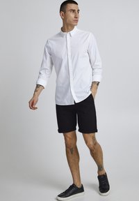 Weißes Hemd mit Knöpfen, schwarze gerollte Shorts und schwarze Sneakers. Glattes Material, lässige Passform und minimalistischer Stil.