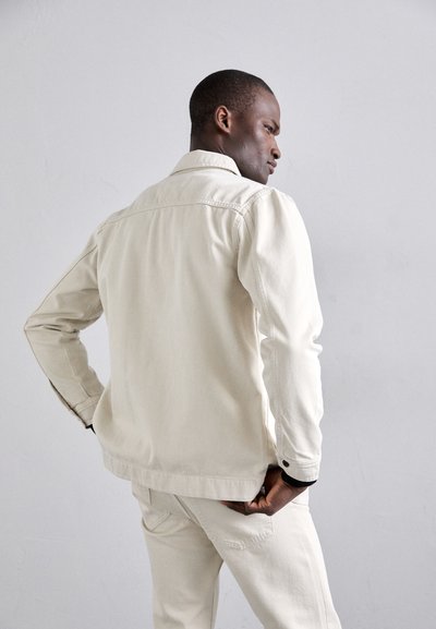 Cream-colored denim jacket with a collared neck, long sleeves, and a relaxed fit, featuring black button accents and a smooth texture.