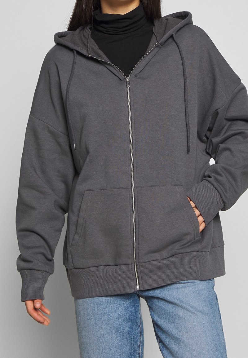 Sudadera gris con cremallera, hecha de un tejido suave, con una capucha grande, bolsillos frontales y puños de canalé. Corte holgado y finalizada con una cremallera.