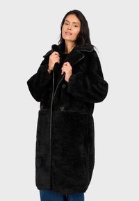 Cappotto nero peloso con un ampio colletto, due grandi bottoni e un tessuto strutturato. Il cappotto ha maniche lunghe a vestibilità comoda e un orlo dritto.