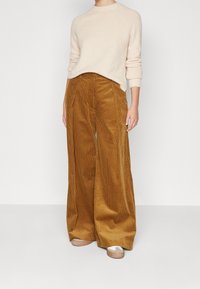 Persona che indossa un maglione lavorato a maglia beige, pantaloni di velluto a coste giallo senape a gamba larga e sneaker beige, in piedi contro uno sfondo bianco semplice.