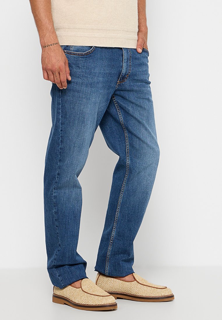 Jasno beżowy sweter, niebieskie dżinsy o prostym kroju oraz beżowe plecione buty na wsuwkę z brązowymi akcentami.