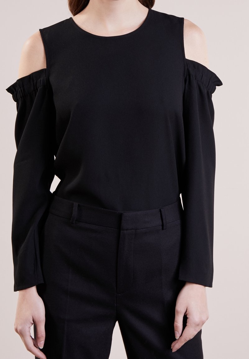 Svart långärmad topp med avskuren axel och volangdetalj, mjuk textur, rund halsringning. Kombinerad med svarta skräddarsydda byxor.