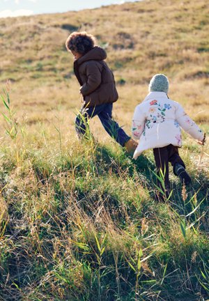 Twee kinderen in winterjassen lopen op een zonnige dag een heuvel op door een grasveld, één draagt een gebreide muts.