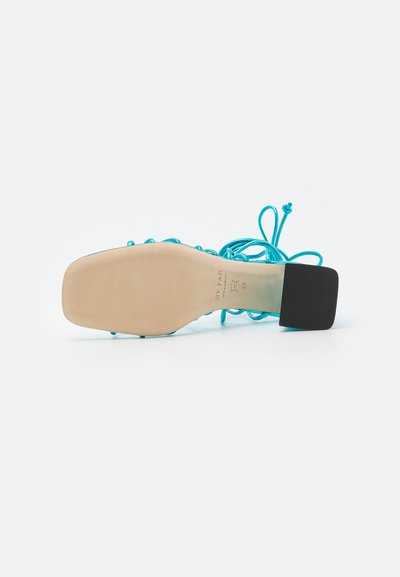 Τυρκουάζ ψηλοτάκουνη σανδάλι με τετράγωνη μαύρη φτέρνα, διαθέτοντας δέσιμο με κορδόνια και ομαλή μπεζ σόλα. Μινιμαλιστικός σχεδιασμός και υφή.