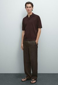 Kortärmad burgundy polo T-shirt i kombination med lösa bruna linnebyxor och svarta flip-flops, stående mot en ljusgrå bakgrund.