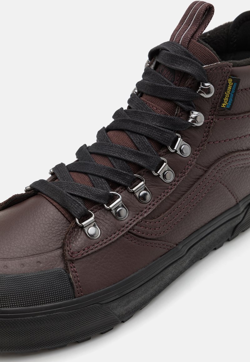 Brązowe skórzane sneakersy za kostkę z czarnymi tekstylnymi sznurowadłami, metalowymi oczkami, strukturalnymi wzorami po bokach oraz gumową podeszwą. Zawierają wodoodporne materiały.