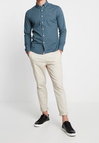 Blå skjorta med lång ärm och knapp, med en liten logotyp, kombinerad med krämfärgade chinos och svarta sneakers. Slät tyg, skräddarsydd passform, uppvikt ärmslut.