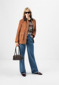 Donna che indossa una giacca di pelle scamosciata marrone, un maglione a fantasia, jeans a palazzo blu, occhiali da sole neri, mentre tiene una borsa marrone scuro, in piedi su uno sfondo bianco.