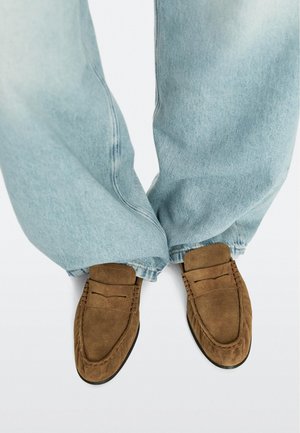 Lyseblå denimjeans båret med brune ruskind-sko på en hvid baggrund, der viser fødder og underben.