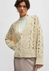 Cardigan crema a maglia con motivi a treccia e pizzo, chiusura con bottoni, polsini a costine e dettagli a trama aperta, indossato sopra una maglietta bianca a collo alto.