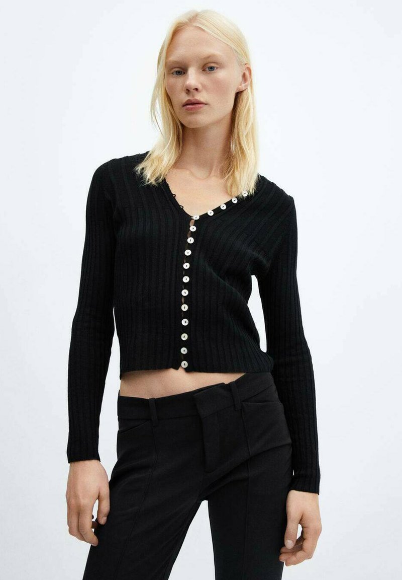 Donna bionda che indossa un cardigan corto nero a coste con bottoni bianchi e pantaloni neri a vita alta su uno sfondo semplice.