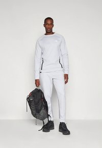 Conjunto atlético cinza claro com mangas compridas e bolsos com fecho, combinado com uma mochila preta com detalhes vermelhos e sapatos pretos resistentes.