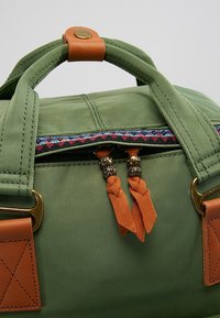 Zelená nylonová taška s hnědými koženými detaily, barevným tkaným lemem a dvojitými zipy s oranžovými třásněmi a dekorativními prvky.