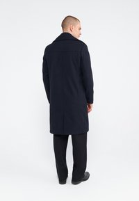 Marineblauwe overjas met een op maat gemaakt ontwerp, met een gekartelde revers en lange mouwen. Gladde stof, tot op de knie, met een enkele rugventilatie.