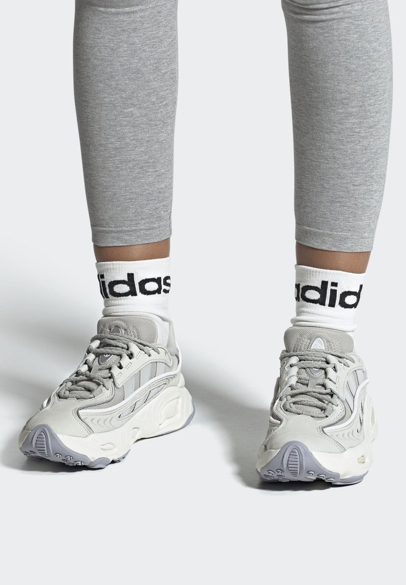 Helle graue Sneakers mit einem strukturierten Design, ausgestattet mit weißen Akzenten, Schnürsenkeln und einer Gummisohle. Kombiniert mit weißen Socken, die das "adidas"-Logo zeigen.