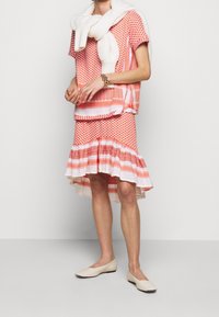 Žena v červeném a bílém vzorovaném topu s krátkým rukávem a odpovídající sukni s asymetrickým lemem, krémových balerínách, s bílým svetrem přehozeným přes ramena a náramkem.