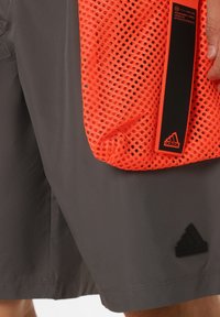 Šedé šortky s matnou textúrou, so čiernym logom Adidas. K nim je pripojená jasne oranžová sieťovaná kapsa s čiernym popruhom.