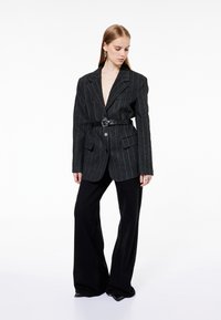 Musta teksturoitu bleiseri syvällä V-aukolla, joka on kiinnitetty leveällä mustalla vyöllä. Yhdistetty mustiin leveälahkeisiin housuihin. Hienovaraisia kuvioita ja kaksi etutaskua.