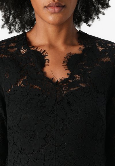 Robe en dentelle noire avec un motif floral, décolleté festonné et détails transparents. Tissu texturé avec des broderies complexes et des accents en tissage ouvert.